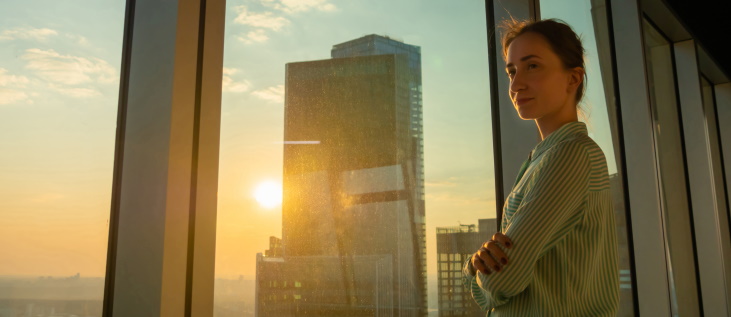 Portrait of smiling woman looking at cityscape through window of skyscraper. Summer time, sunset, lens flare. Relax, leisure time, success, opportunity, sightseeing, discover and future concept